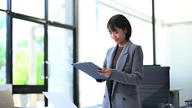 Asian businesswoman in blazer reviewing report on clipboard while standing at desk. Confident, professional, strategic leader in modern office.