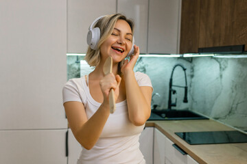 Happy young woman singing with wooden spoon like microphone, wearing headphones in modern kitchen. Joyful moment, music and fun at home.