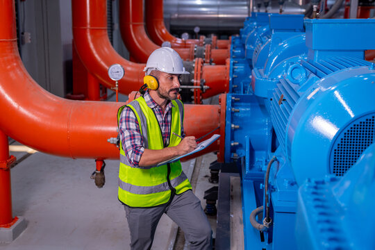 Engineers wearing safety helmets and reflective vests inspect large industrial machinery inside a factory check pressure gauges, take notes clipboard maintenance workplace system operation and safety.