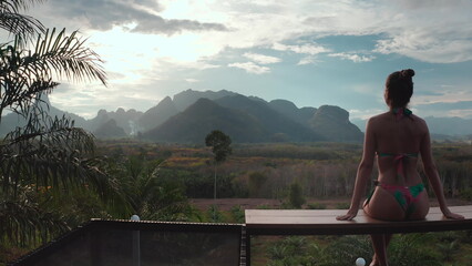 Woman in bikini sit on deck looking stunning view sunset mountain valley in Thailand resort. Girl relax in holiday exotic wild nature hut villa. Outdoor lifestyle travel on summer vacation back view