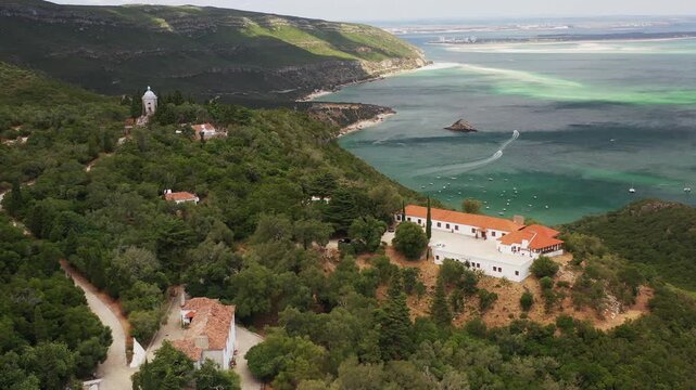 Drone footage of Convento de Nossa Senhora da Arrabida nestled in the forested hills of Arrabida Natural Park, Portugal, showing scenic architecture surrounded by lush green landscape