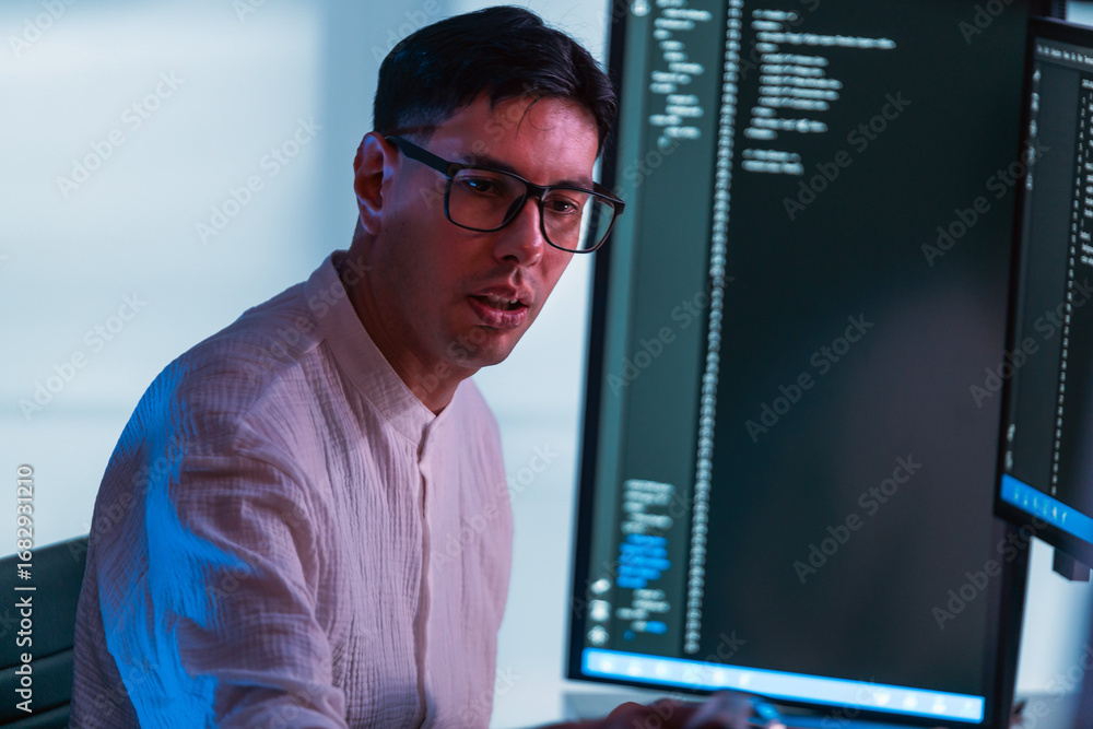 Poster programmer wearing glasses codes at dual monitors in modern office, lit by cool blue light. scene hi