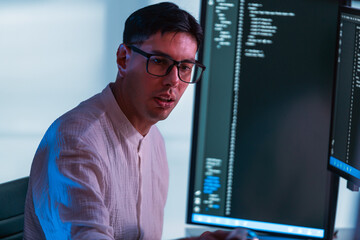 Programmer wearing glasses codes at dual monitors in modern office, lit by cool blue light. Scene...