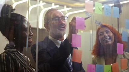 Diverse business team in modern office brainstorms as senior manager writes on glass with sticky notes. Corporate meeting showcases teamwork, collaboration, leadership, engaged colleagues. 4k video