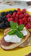 Pancakes with sour cream, blackberries, raspberries, and mint leaves on a vibrant yellow plate