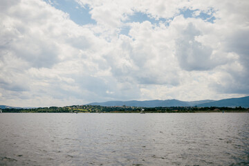 Calm lake water reflecting cloudy sky, showcasing distant mountains and charming lakeside village. Sailboats gracefully navigating serene landscape on warm summer day. Tranquil alpine lake