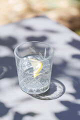 Single glass of water with lemon slice on outdoor table in filtered sunlight with dramatic shadow patterns