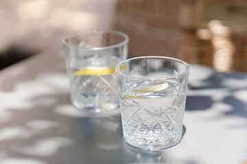 Two crystal glasses with lemon-infused water on a gray table in dappled sunlight, casting soft...