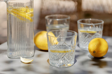 Glass pitcher with lemon water, faceted glasses, whole lemons and a lemon wedge on a shaded outdoor...