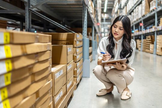 Focused on her task, young asian woman sitting and holding clipboard meticulously updates inventory records while surrounded by shelves filled with various products in a vibrant warehouse setting - Powered by Adobe