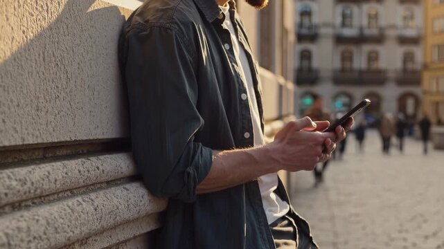 A man stands in the sunlight, using his phone while leaning against a wall. The man is focused on his phone, creating a sense of detachment from the surrounding city scene.