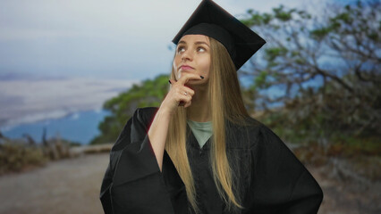 Woman pondering in graduation cap and gown outdoors, with nature background symbolizing future possibilities and achievement.