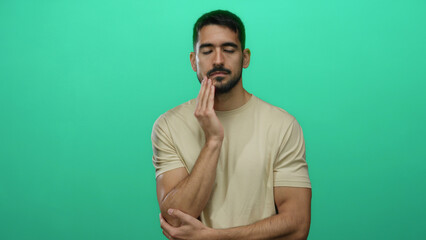 Young hispanic man with beard in beige shirt pondering thoughtfully against vibrant green background with arms crossed and eyes closed