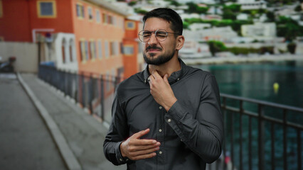 Young man in glasses and gray shirt stands on promenade near seaside holding throat with distressed...