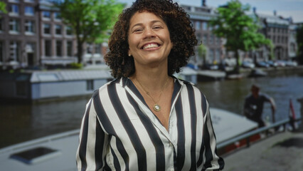 Woman with curly hair sticks her bare tongue out and smiles on a canal boat on a street;...