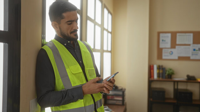 Hispanic man wearing vest holds phone looking thoughtful in bright office leaning against window exuding contemplation.
