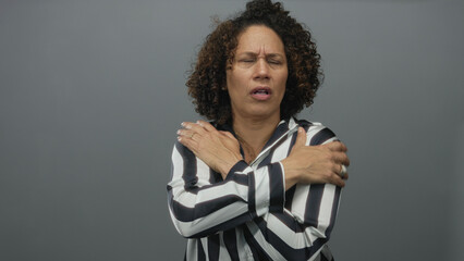 Naklejka premium Middle aged hispanic woman arms crossed hugging chest in gray studio wearing black and white striped blouse and rings, tense expression; cold discomfort.