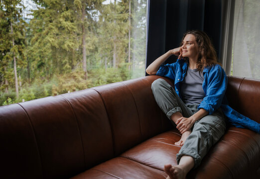 Young smiling woman in blue shirt sitting on sofa at home looking at forest outside window - Powered by Adobe