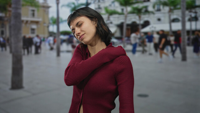 Young hispanic woman in burgundy sweater grimaces and holds shoulder for pain while standing on street; discomfort.