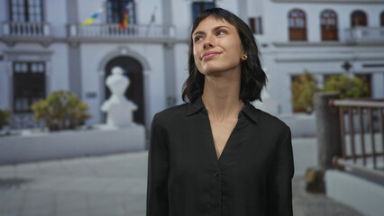 Young hispanic woman tilts head and smiles gently against white building, dark hair framing face;...