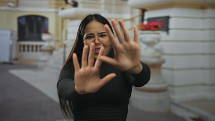 Fototapeta premium Woman with glasses gesturing stop in an urban outdoor setting showcasing expression with a blurred historic building background, emphasizing emotion and street environment.