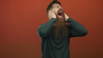Young caucasian man with long beard shouting with hands on ears in studio with red background;...