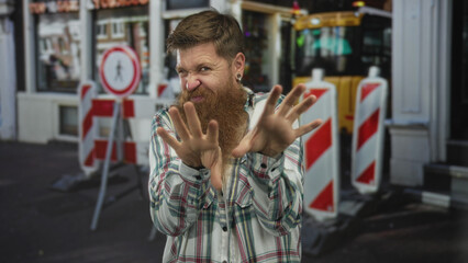 Man pinches nose with both hands while scrunching face in front of traffic barriers on colorful urban street; disgust.