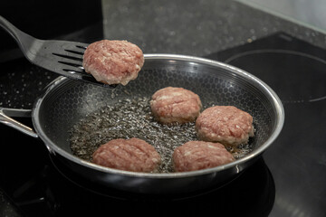 Fresh raw cutlets sizzling in hot oil within a pan on a modern induction cooktop, with a spatula lifting one, symbolizing home meal preparation