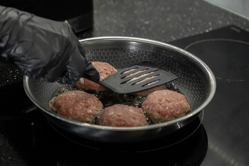Person in black glove frying raw cutlets with a spatula in a hot pan on an induction stove for home cooking, healthy meal preparation