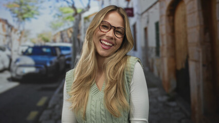 Fototapeta premium Woman wearing glasses and a green sweater vest smiles with eyes closed on a sunlit cobblestone street lined with parked cars; happiness contentment.