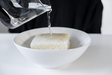 Person wearing black glove pours clear fresh water onto white toast bread slices in ceramic bowl, showcasing liquid absorption for culinary or scientific use