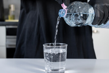 Person in black protective gloves pouring refreshing clean water from a plastic bottle into a clear...