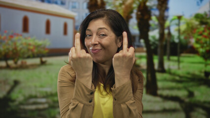 Middle age hispanic woman smirking flips both middle fingers in sunlit green park wearing brown jacket and yellow shirt; defiance.