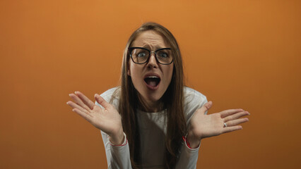 Woman with raised palms, open mouth and glasses in orange studio showing a startled shrug and wide...