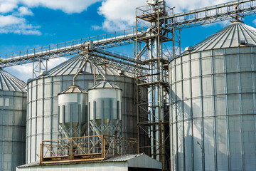 Multiple large metal grain silos with intricate conveyor systems under a bright blue sky. Modern...