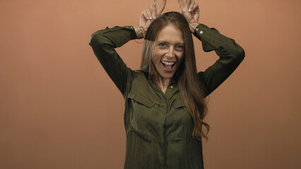 Woman making bunny ears with fingers and smiling in studio with peach backdrop wearing olive shirt;...