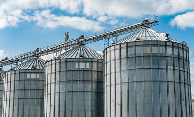 Large industrial metal grain silos facility with conveyor system for agricultural storage under blue sky. © SMK