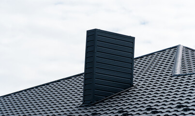 Dark grey modern tiled roof and rectangular chimney against an overcast sky. Architectural detail.