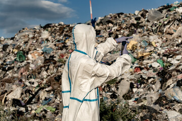 Person in protective suit pointing device at massive garbage landfill. Environmental waste...