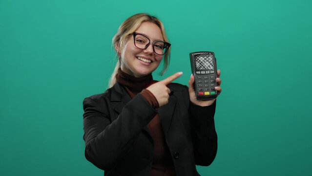 Woman smiling holding a payment terminal in a business suit pointing forward on a bright green background highlighting banking technology and customer interaction