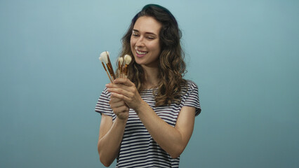 Woman holds makeup brushes and smiles in studio against a light blue wall, wearing a striped shirt and showcasing curly hair; beauty creativity joy.