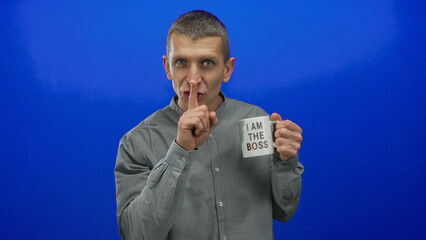 Caucasian man in a gray shirt gesturing silence and winking while holding a coffee cup labeled 'i am the boss' against an isolated blue wall background.