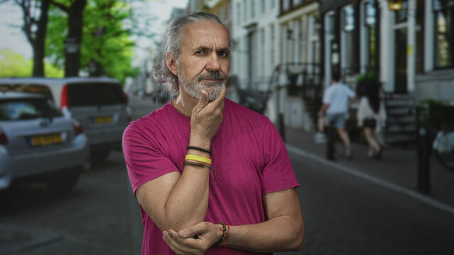 Man with hand on chin and wrist bracelets in magenta shirt standing on a street lined with parked cars and townhouses; introspection. - Powered by Adobe