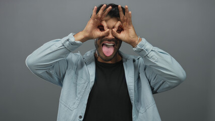 Hispanic man with short black hair making binocular gesture with hands and showing tongue in grey...
