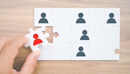 Hand placing a red figure jigsaw piece in a puzzle with gray figures on wood. Symbolizes team, unique talent, or recruitment