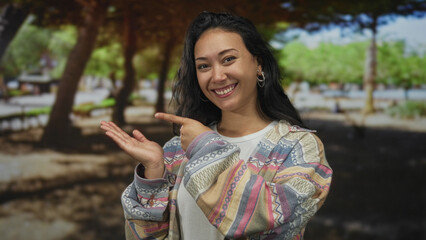 Woman smiling and pointing finger at open palm in forest wearing patterned sweater and hoop earring; friendly greeting joy.