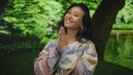 Fototapeta premium Woman with palms pressed together near chest in green park by a tree, smiling warmly and looking at viewer; joy gratitude.