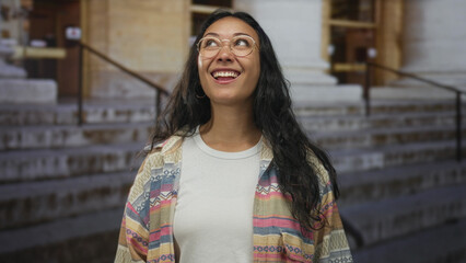 Woman wearing glasses smiles and looks up on building steps with patterned jacket and white tshirt visible; carefree joy.