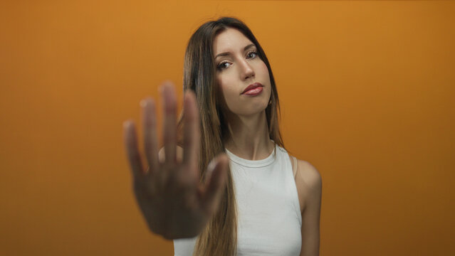 Confident hispanic woman wearing white tank top extends palm forward in studio with orange wall; defiance.