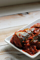 Sarden or Sardencis. Spicy Indonesian mackerel dish served in rich red chili sauce with lemongrass on a white plate over rustic wooden table.